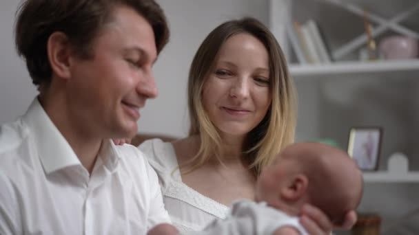 Rack focus from happy Caucasian mother putting head on husband shoulder to smiling father admiring cute baby son. Loving husband and wife enjoying weekend leisure with newborn child indoors at home. — ストック動画