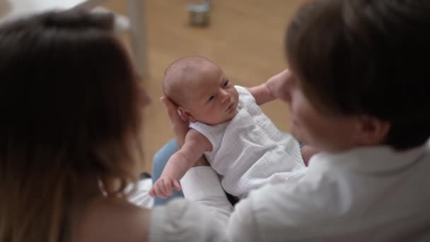 Portret van een schattige pasgeboren jongen met een paar ouders thuis in de woonkamer. Schieten over de schouder van vader en moeder bewonderende schattige blanke zuigeling zittend op de bank binnen. Langzame beweging. — Stockvideo