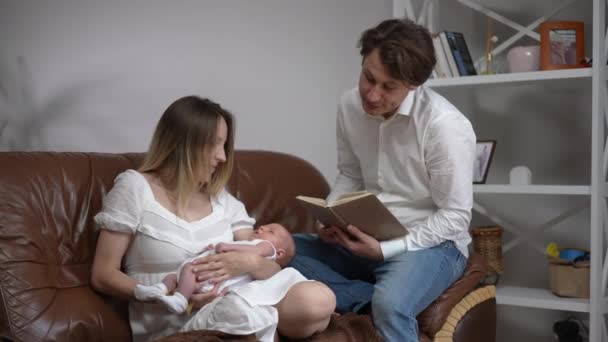 Portrait of smiling father singing lullaby reading book sitting with wife and newborn son on couch at home. Cheerful Caucasian man enjoying infant nap time with woman and baby boy in slow motion. — Video