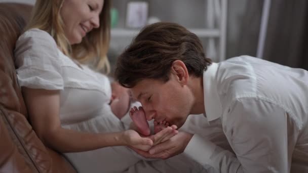 Happy smiling father kissing tiny baby toes of newborn child in hand of mother. Side view portrait of Caucasian smiling man admiring small feet of little infant son at home indoors. Slow motion. — Stok Video