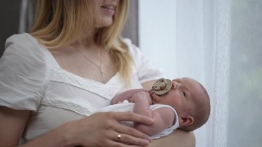Calm Caucasian newborn boy sucking pacifier in slow motion lying on hands of unrecognizable woman. Portrait of cute little infant baby enjoying leisure with mother indoors at home. Tranquility.
