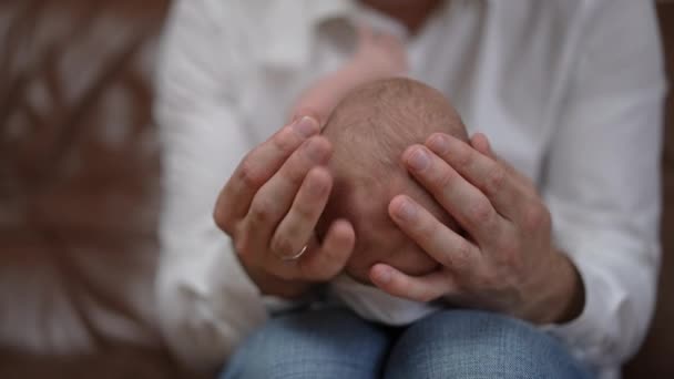 Close-up head of newborn baby in male Caucasian hands caressing child. Unrecognizable father enjoying leisure with infant son indoors at home sitting on couch. Slow motion. — Wideo stockowe