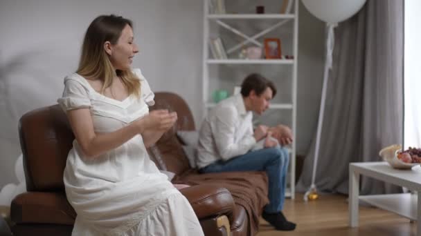 Loving happy Caucasian woman admiring man and newborn infant sitting at background looking at camera gesturing in slow motion touching chest. Concept of family love and happiness. — Stock Video