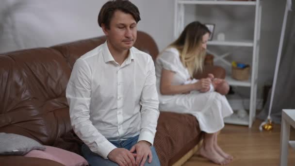 Thankful happy father husband admiring woman and newborn son sitting at background turning to camera holding hands together. Portrait of happy Caucasian man posing indoors with wife and infant child. — Stock video