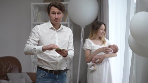 Caucasian man scattering money in slow motion with woman and newborn infant standing at background. Portrait of upset husband father spending lot of cash on family posing indoors at home. — стоковое видео