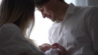 Mutlu gülümseyen baba, yeni doğan oğlunun annesinin elinde ailesiyle pencerenin önünde durmasına hayran. Beyaz adamın portresi evde karısı ve bebeğiyle sakin bir şekilde vakit geçiriyor. Yavaş çekim.