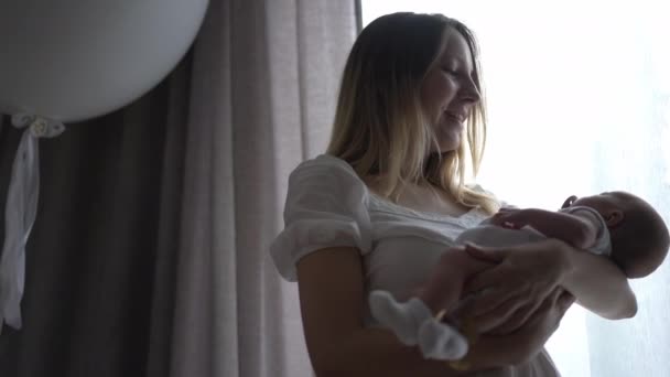 Портрет коханої кавказької жінки з новонародженою дитиною в руках стоїть біля вікна в приміщенні, милуючись немовлям. Щаслива приваблива мати проводить дозвілля з маленьким сином вдома. Повільний рух. — стокове відео