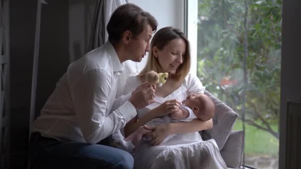 Loving father playing toys talking to newborn son in hands of woman sitting on windowsill. Happy Caucasian couple of parents admiring little infant enjoying leisure indoors in slow motion. Family joy. — Video Stock