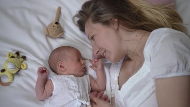 Happy woman talking with newborn boy lying on comfortable soft white bed at home. Top view portrait of excited Caucasian mother admiring infant baby kissing tiny hand in slow motion. — Stockvideo