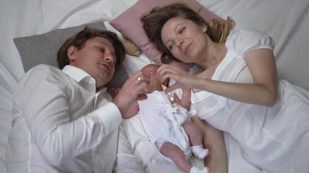 Happy couple gesturing heart shape with hands lying on bed with newborn baby at home indoors looking at camera. Confident Caucasian husband and wife posing with infant son in slow motion. — Video Stock