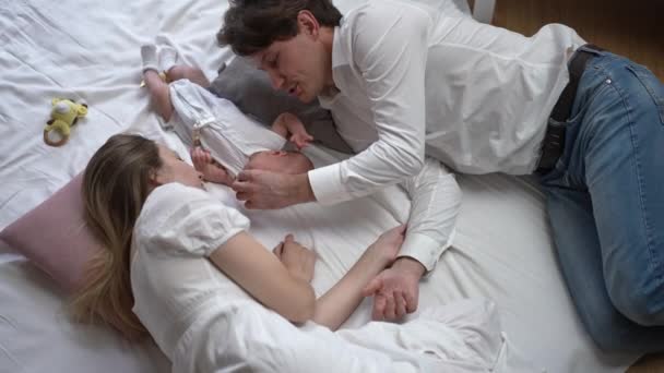 Κάτοψη άντρας και γυναίκα ξαπλωμένοι με νεογέννητο αγόρι σε λευκό μαλακό κρεβάτι στο σπίτι αναπαύονται. Ευτυχισμένος Καυκάσιος πατέρας μητέρα και το βρέφος γιος απολαμβάνουν αναψυχής σε εσωτερικούς χώρους. Αργή κίνηση. — Αρχείο Βίντεο
