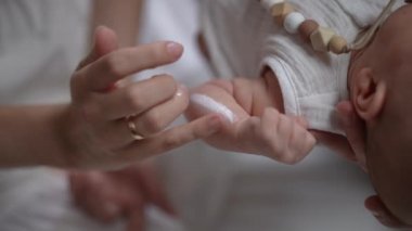 Close-up female finger rubbing moisturizer in hand of newborn baby in slow motion. Unrecognizable Caucasian mother taking care of infant son at home. Child care and love concept.