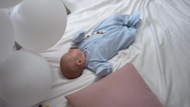 Wide shot top view calm newborn boy in blue lying on cozy bed with balloons sucking pacifier falling asleep. Happy carefree Caucasian infant baby sleeping at nap time indoors at home. Slow motion. — Vídeo de stock