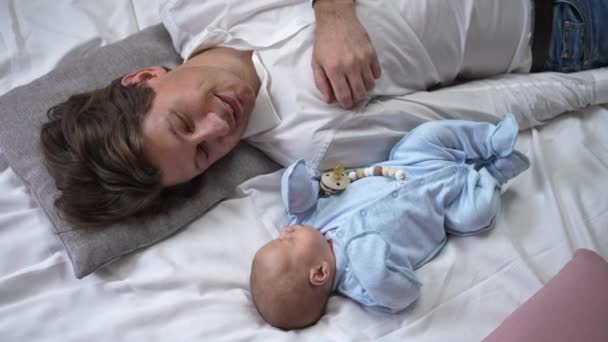 Smiling Caucasian man admiring newborn son in blue lying on comfortable cozy bed with baby indoors. Portrait of loving father resting with infant kid enjoying leisure. Slow motion. — 图库视频影像