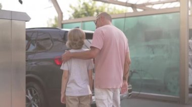 Arkadan bakan adam, dışarıda araba yıkama servisinde duran temiz aracın önünde konuşan çocuğu kucaklıyor. Yazın oğluyla birlikte araba temizleyen dövmeli beyaz bir baba. Yavaş çekim.