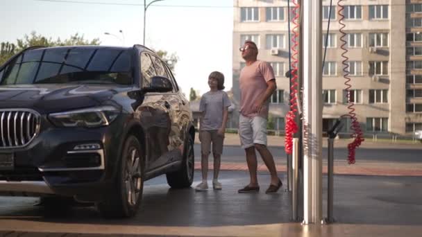 Weitwinkelporträt eines kaukasischen Mannes und Jungen, die in Zeitlupe an einem sauberen Auto in einer Waschanlage reden. Selbstbewusste Vater-Sohn-Reinigung im Freien an einem Sommertag. Autopflege. — Stockvideo