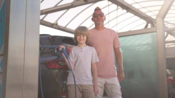 Μέτριο πλάνο πορτρέτο του ικανοποιημένου πατέρα και του γιου στέκεται στη λιακάδα στο πλυντήριο αυτοκινήτων κοιτάζοντας κάμερα χαμογελώντας. Θετικός Καυκάσιος άνδρας και αγόρι ποζάρουν σε αργή κίνηση έξω την ηλιόλουστη μέρα. — Αρχείο Βίντεο