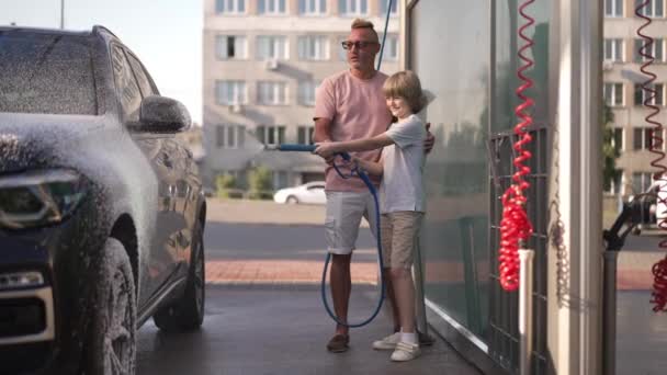 Buon figlio che aiuta il padre a lavare il veicolo all'autolavaggio all'aperto sorridendo. Positivo ragazzo caucasico con rondella ad alta pressione spruzzando schiuma sull'automobile con l'uomo che parla bambino di supporto. — Video Stock