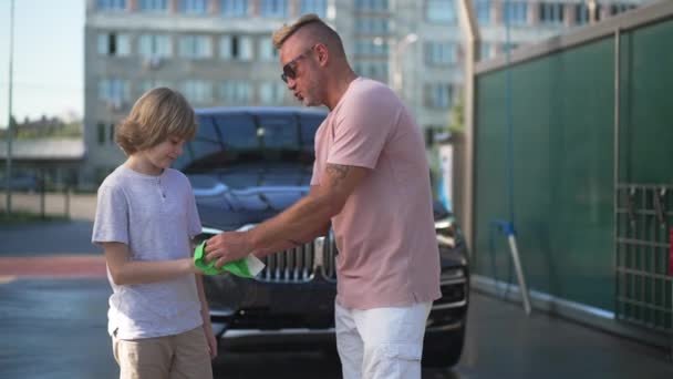 誇り高い白人の父親の息子と車の洗浄サービスの子供の手を布でこすりできれいな車に立って話しています。車を洗うのを手伝ってくれた少年に感謝する幸せな入れ墨の男。スローモーション. — ストック動画