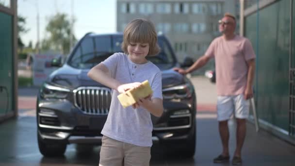 Veselý roztomilý chlapec stlačování vody z houby na mytí aut služby stojící v čistém automobilu s rozmazaný muž v pozadí. Přední pohled portrét šťastný syn pomáhá otci mytí vozidla. — Stock video