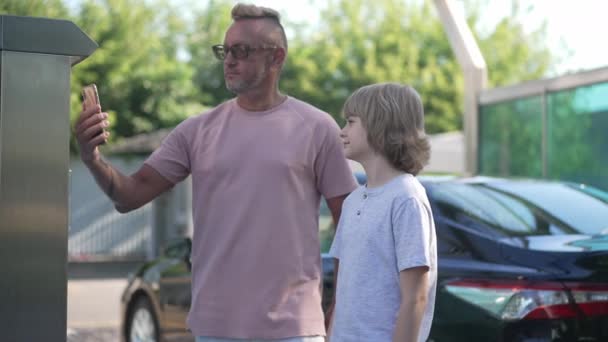 Επικεντρωμένος Καυκάσιος που πληρώνει για πλύσιμο αυτοκινήτων με smartphone σε εξωτερικούς χώρους με το γιο του σε μαύρο όχημα. Συγκεντρωμένος επιτυχημένος πατέρας και γιος στο πλυντήριο αυτοκινήτων. Αργή κίνηση. — Αρχείο Βίντεο