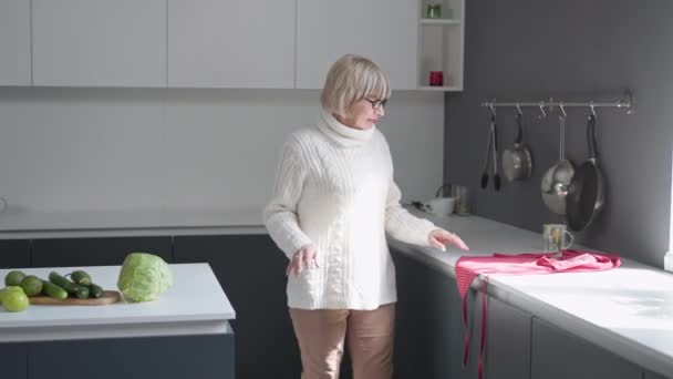 Elegante donna caucasica anziana che cammina in cucina soleggiata indossando grembiule sorridente. Bella pensionato fiducioso in occhiali da vista cucinare sano cibo fatto in casa al chiuso a casa al mattino. — Video Stock