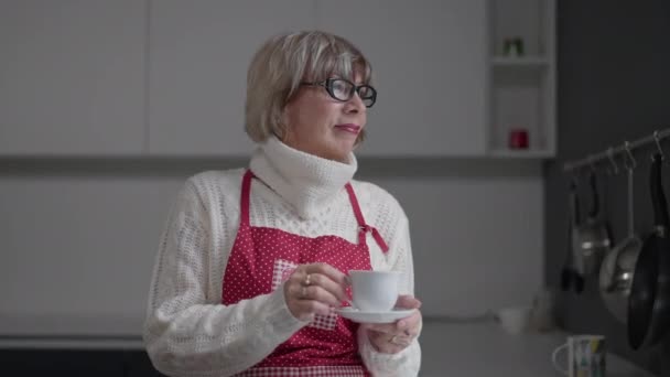 Senior mujer sonriente reflexiva de pie con taza de café en la cocina mirando hacia otro lado. Retrato de mujer caucásica atractiva positiva jubilada disfrutando de la mañana mirando por la ventana. — Vídeo de stock