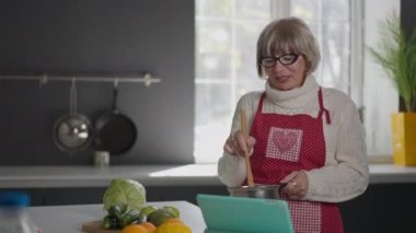 Kıdemli kadın tablette gülen video blogu hakkında konuşan malzemeleri karıştırıyor. Kendinden emin beyaz blogcu emeklinin portresi mutfakta lezzetli ev yapımı yemekler pişiriyor..