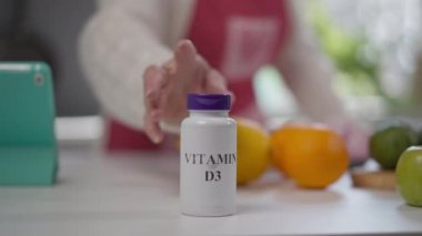 Yakın plan D3 vitamini şişesi masada, son sınıf beyaz eli kavanozu kaldırıyor. Tanımlanamayan emekli kadın evde ilaç alıyor. Sağlık ve yaşam tarzı kavramı.