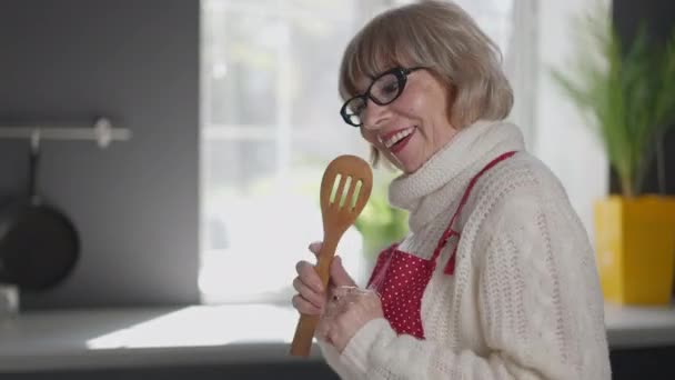 Glædelig kaukasiske kvindelige pensionist have det sjovt at danse sang i køkkenet derhjemme indendørs. Positiv smuk senior kvinde nyder madlavning middag og hobby. Aktiv livsstil koncept. – Stock-video