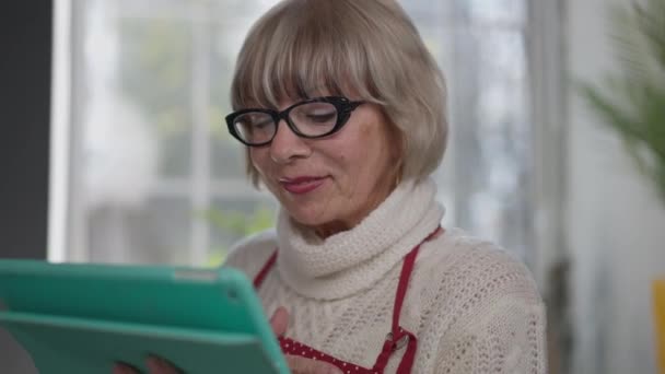 Дуже щаслива жінка на пенсії говорить посміхаючись, використовуючи відео чат на планшеті в приміщенні. Портрет чарівної кавказької старшої жінки в окулярах, яка насолоджується бездротовим спілкуванням вдома.. — стокове відео