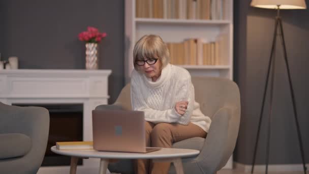 Breed shot portret van drukke blanke senior vrouw surfen op internet op laptop zittend op fauteuil in de woonkamer. Geconcentreerde gepensioneerde berichten online binnen. Gemak en comfort. — Stockvideo