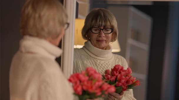 Reflejo en espejo de mujer mayor con ramo de flores de pie en el interior. Hermosa mujer caucásica jubilada admirando tulipanes rojos en la sala de estar en casa. Cumpleaños o celebración del Día de la Mujer. — Vídeos de Stock