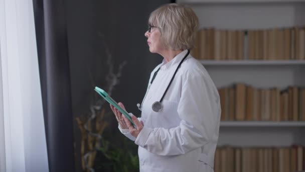 생각있는 나이든 여성 의사가 창 밖을 내다보며 태블릿으로 인터넷 서핑을 한다. 안경을 쓰고 흰 가운을 입고 있는 문제를 겪고 있는 백인 여자의 모습. — 비디오
