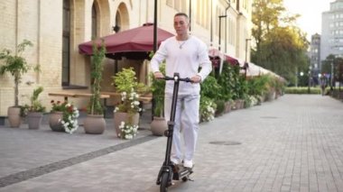 Şehir caddesinde ağır çekimde elektrikli scooter kullanan geniş açılı bir milenyum adamı. Yaz günü şehirde e-scooter kullanan kendine güvenen yakışıklı beyaz bir adamın portresi..