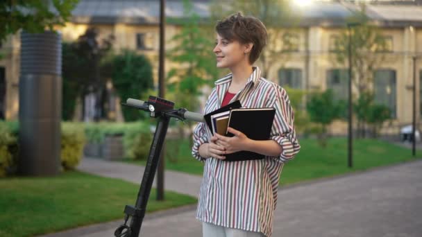 Jeune femme aux cheveux courts debout sur la rue de la ville avec scooter électrique et des livres regardant autour et regardant la caméra souriant. Portrait de confiant intelligent femme caucasienne étudiant posant à l'extérieur. — Video