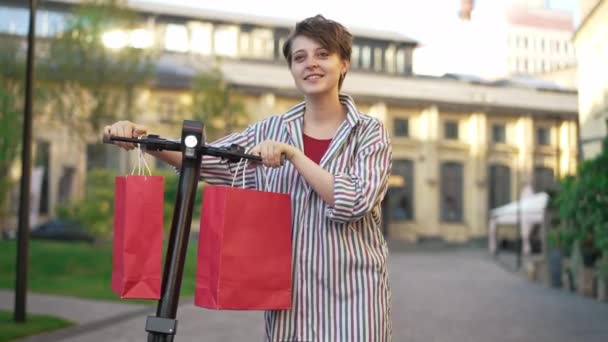 Přední pohled jistý okouzlující mladá žena chůze s e-scooter a nákupní tašky visí na volantu. Portrét spokojeného kavkazského nakupujícího s nákupy ve zpomaleném filmu. — Stock video