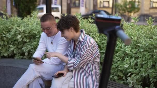 Vidám pár, akik nevetve szörföznek a közösségi médiában, a városi utcában ülve, elmosódott e-robogós kormánykerékkel az elején. Boldog gondtalan kaukázusi barát és barátnő szórakozás társkereső. — Stock videók