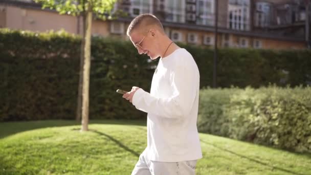 Side view hymyilevä onnellinen mies aurinkolasit kävely auringonsäteet ulkona messaging verkossa älypuhelimella. Rento luottavainen valkoihoinen komea kaveri kävelee kaupungin kadulla tekstiviestejä puhelimessa hidastettuna. — kuvapankkivideo