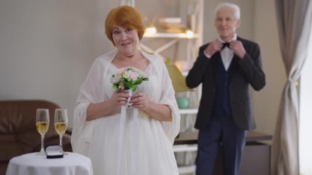 背景に立ってエレガントなスーツでぼやけた新郎としてブライダル花束でポーズ結婚式のドレスで幸せな白人のシニア赤毛の花嫁の肖像画。フロントビューカメラを見て楽しい女性笑顔. — ストック動画
