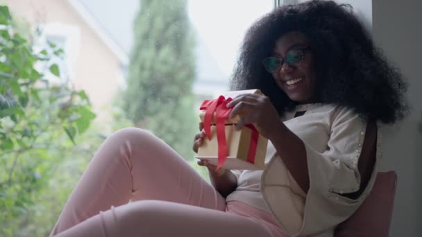 Εκπλήσσομαι που μια Αφρο-Αμερικανίδα ανοίγει κουτί δώρου καθισμένη στο περβάζι χαμογελώντας. Πορτρέτο της χαρούμενης νεαρής όμορφης χιλιετίας κυρία απολαμβάνοντας έκπληξη μέσα στο σπίτι. Αργή κίνηση. — Αρχείο Βίντεο
