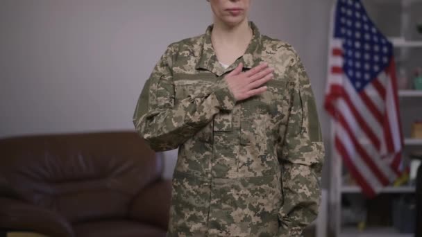 Unrecognizable serious military woman in camouflage uniform putting hand on chest standing with USA flag at background. Confident courageous brave female soldier indoors. Slow motion. — Stock Video