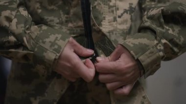 Canlı kamera, kadın elleri ağır çekimde kamuflaj üniforması giyerken ve ciddi bir yüz ifadesiyle kameraya bakarken izliyor. İçeride poz veren kendine güvenen genç kadın portresi..