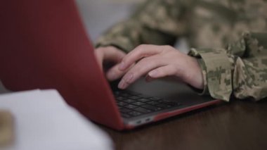 Kadın eli kamuflajlı, dizüstü bilgisayarda ağır çekimde klavyede daktilo kullanıyor. Tanımlanamayan Kafkasyalı askeri üniformalı genç bir kadın internet mesajlarında geziniyor kapalı bir masada oturuyor..