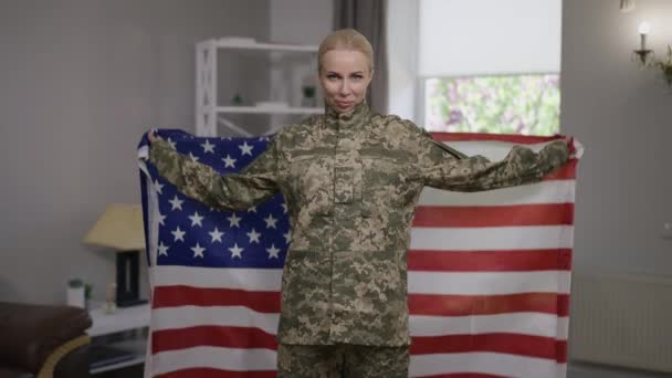 Medelhög bild av positiv säker militär kvinna insvept i amerikanska flaggan leende tittar på kameran. Porträtt av vacker kvinnlig rekryt poserar inomhus i slow motion. — Stockvideo