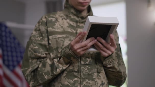 Mulher triste irreconhecível em camuflagem uniforme abraçando imagem na foto moldura família desaparecida. Jovem chateado recrutar feminino recordando memórias em pé dentro de casa com bandeira dos EUA no fundo. — Vídeo de Stock