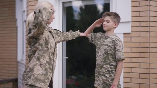 Мальчик-подросток учится отдавать честь в замедленной съемке, когда военная женщина регулирует осанку и руку. Портрет гордых кавказских сыновей, наслаждающихся выходными с матерью на открытом воздухе. Концепция поддержки семьи. — стоковое видео