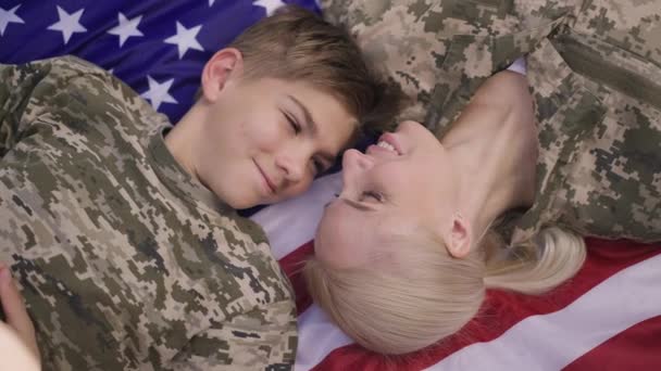 Close-up top view happy relaxed Kaukasische tienerjongen en jonge vrouw in kaki liggend op Amerikaanse vlag lachend en sluitend. Militaire moeder en trotse zoon genieten van picknick buiten. — Stockvideo