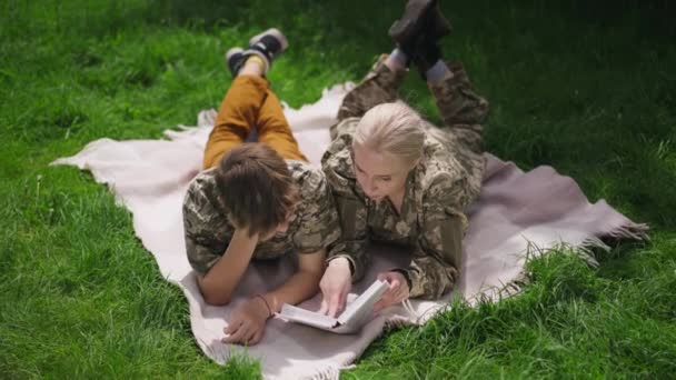 Aus der Vogelperspektive entspannte kaukasische Militärfrau in Tarnkleidung und Teenager-Junge, der auf einer Decke im sonnigen Park beim Lesen eines Buches liegt. Glückliche Mutter und Sohn genießen Hobby am Wochenende im Freien. — Stockvideo