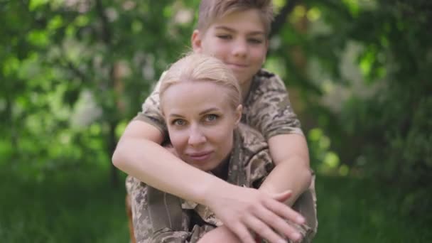 Красивая военная мать смотрит в камеру, улыбаясь, как веселый сын-подросток, обнимающий родителя в замедленной съемке. Увеличить масштаб камеры в замедленной съемке. Счастливая женщина, наслаждающаяся выходными с мальчиком. — стоковое видео